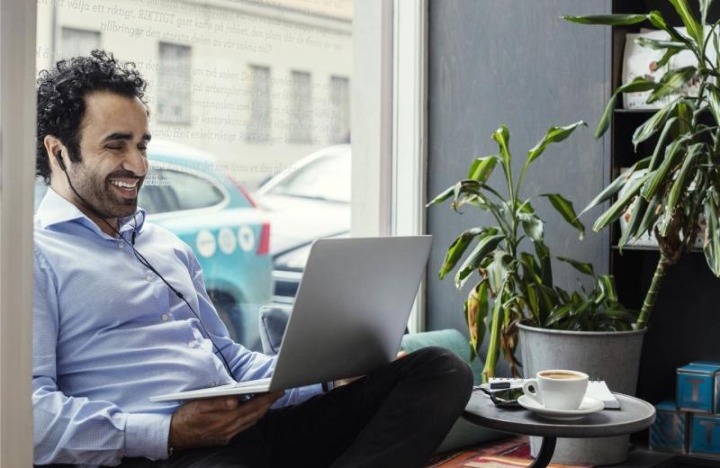 Man sitting on a seat by a window, smiling at a laptop sitting on his lap